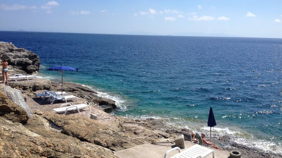 A rocky coastline with clear blue sea, white lounge chairs, and a few people by the water.