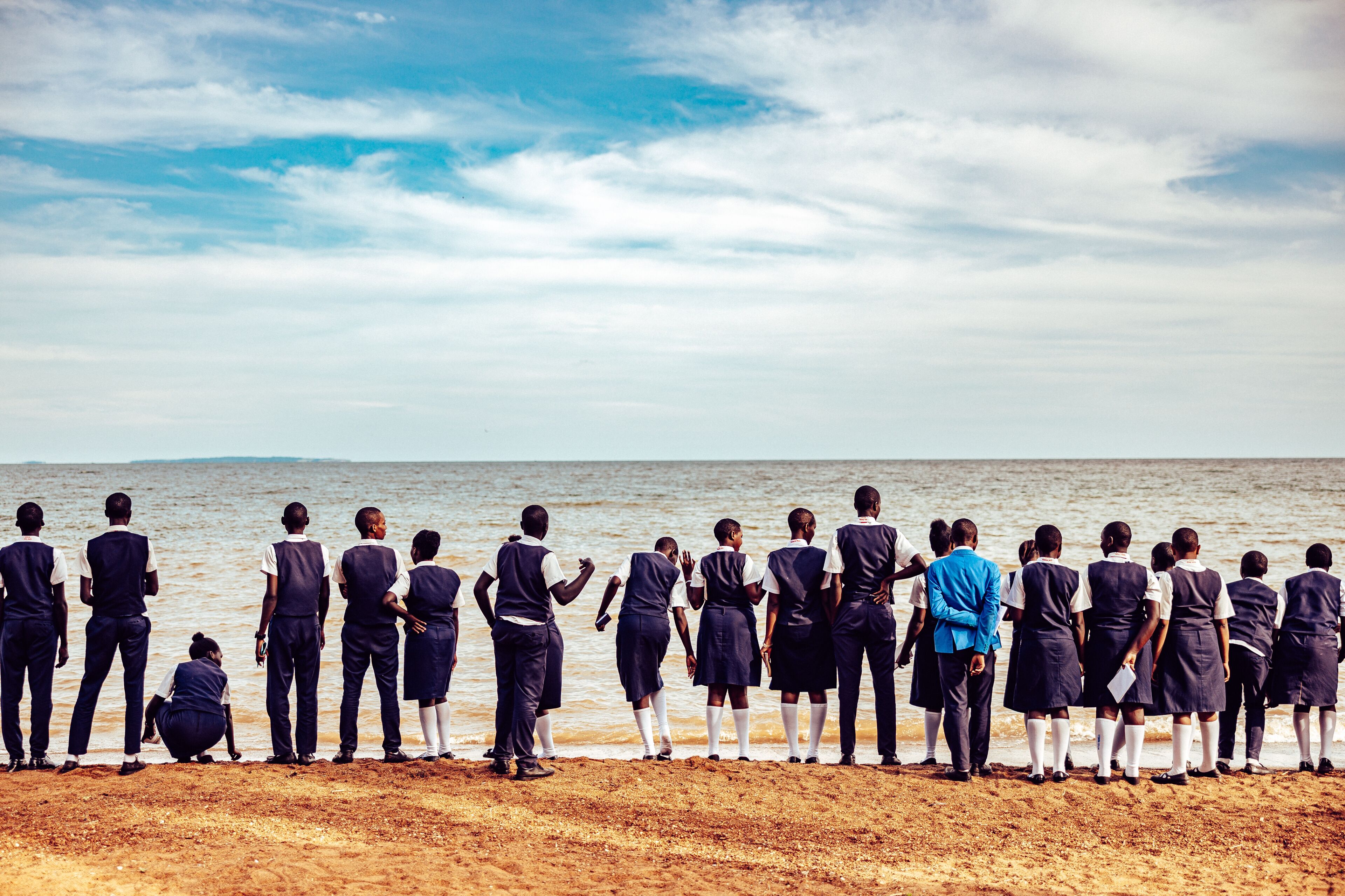 Kampala, Uganda - 26 March 2024: View of students lined up on the sandy beach, gazing out at the serene water meeting the horizon under a partly cloudy sky.