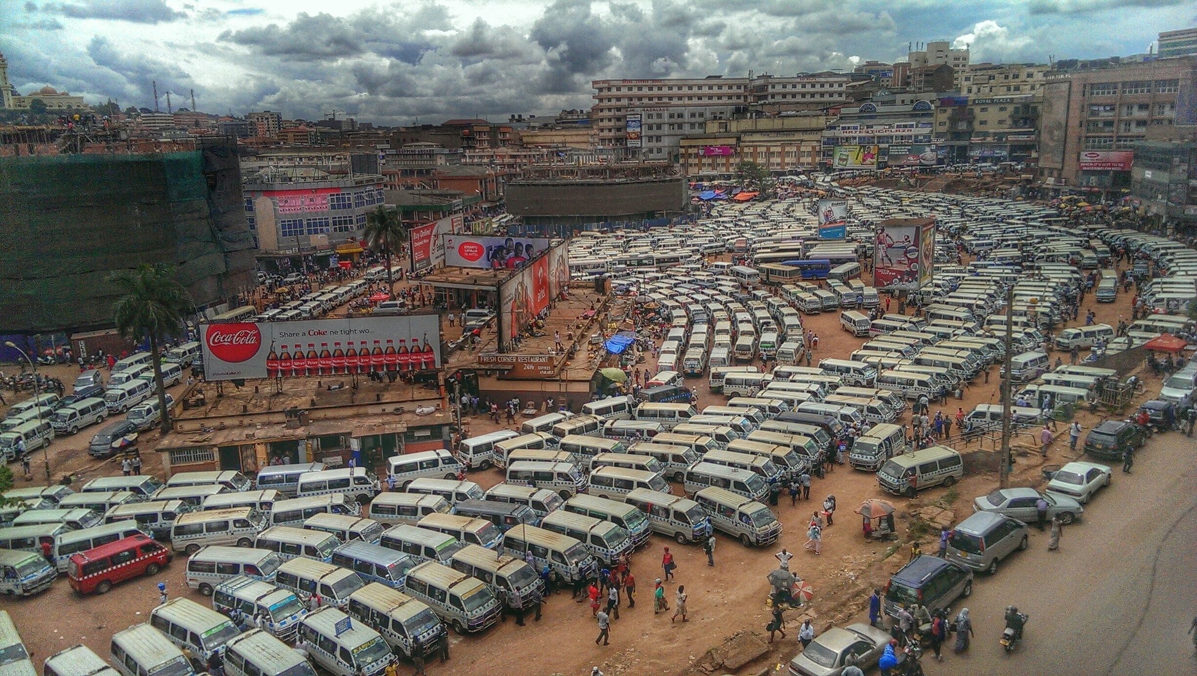 A congested parking lot with numerous buses and cars, a Coca-Cola billboard, and a crowded street scene.