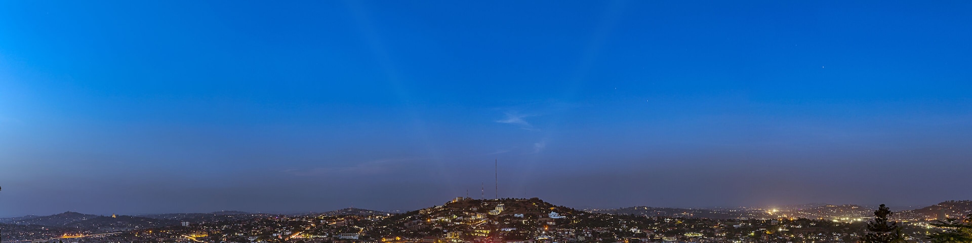 A cityscape at dusk with a prominent hill in the foreground and a clear sky.