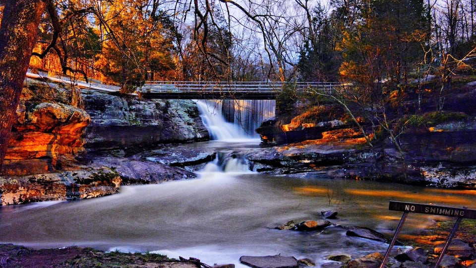 A waterfall, a bridge, and a 'No Swimming' sign in a forested area.