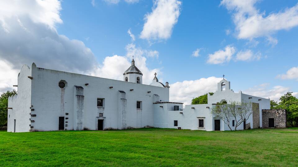 A white colonial-style building with a bell tower and a small chapel, surrounded by green grass and trees.
