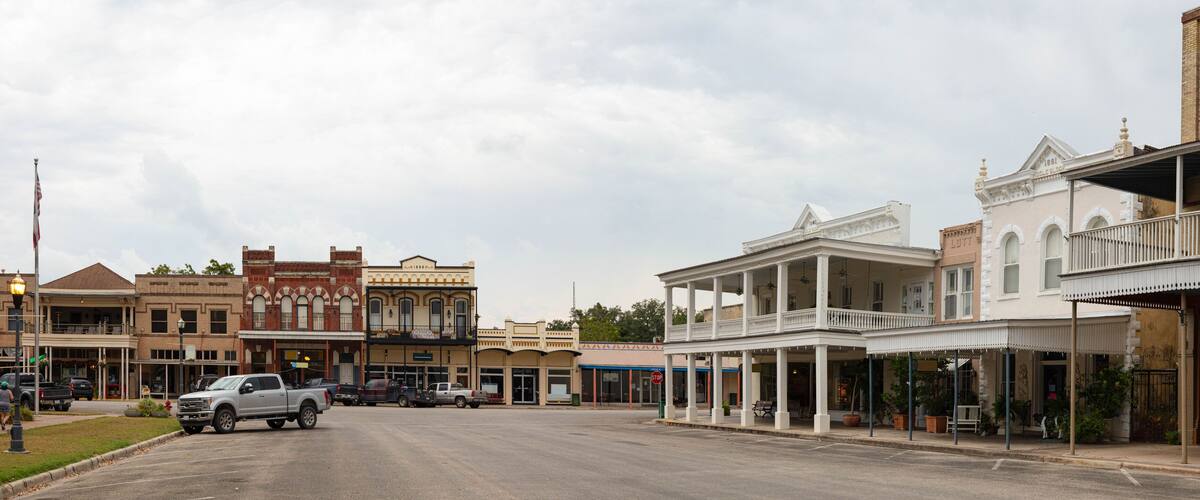 A historic downtown street with vintage buildings, a prominent American flag, and parked vehicles.