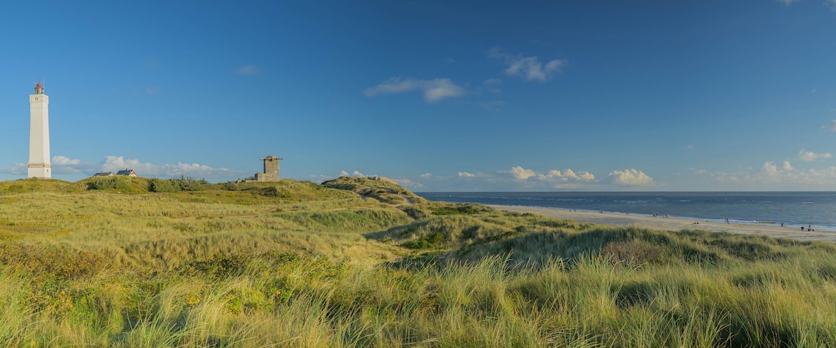 A lighthouse on a grassy hill overlooking a beach with people in the distance.