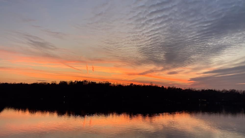 Un coucher de soleil sur un lac paisible, avec la silhouette d’arbres et un ciel nuageux.