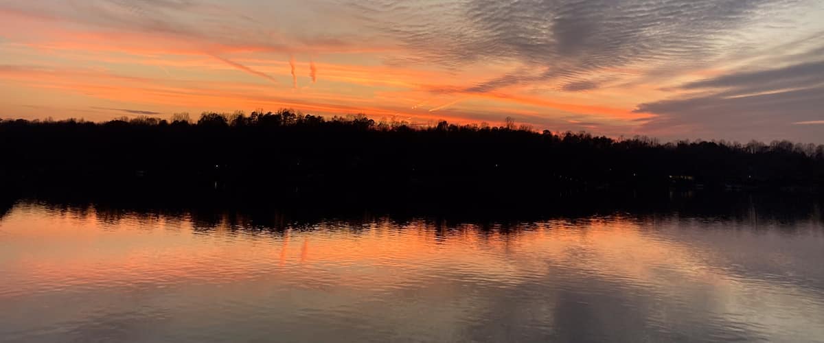 A sunset over a calm lake with a silhouette of trees and a cloudy sky.