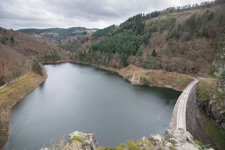 A large reservoir with a dam, surrounded by forested hills and a cloudy sky.