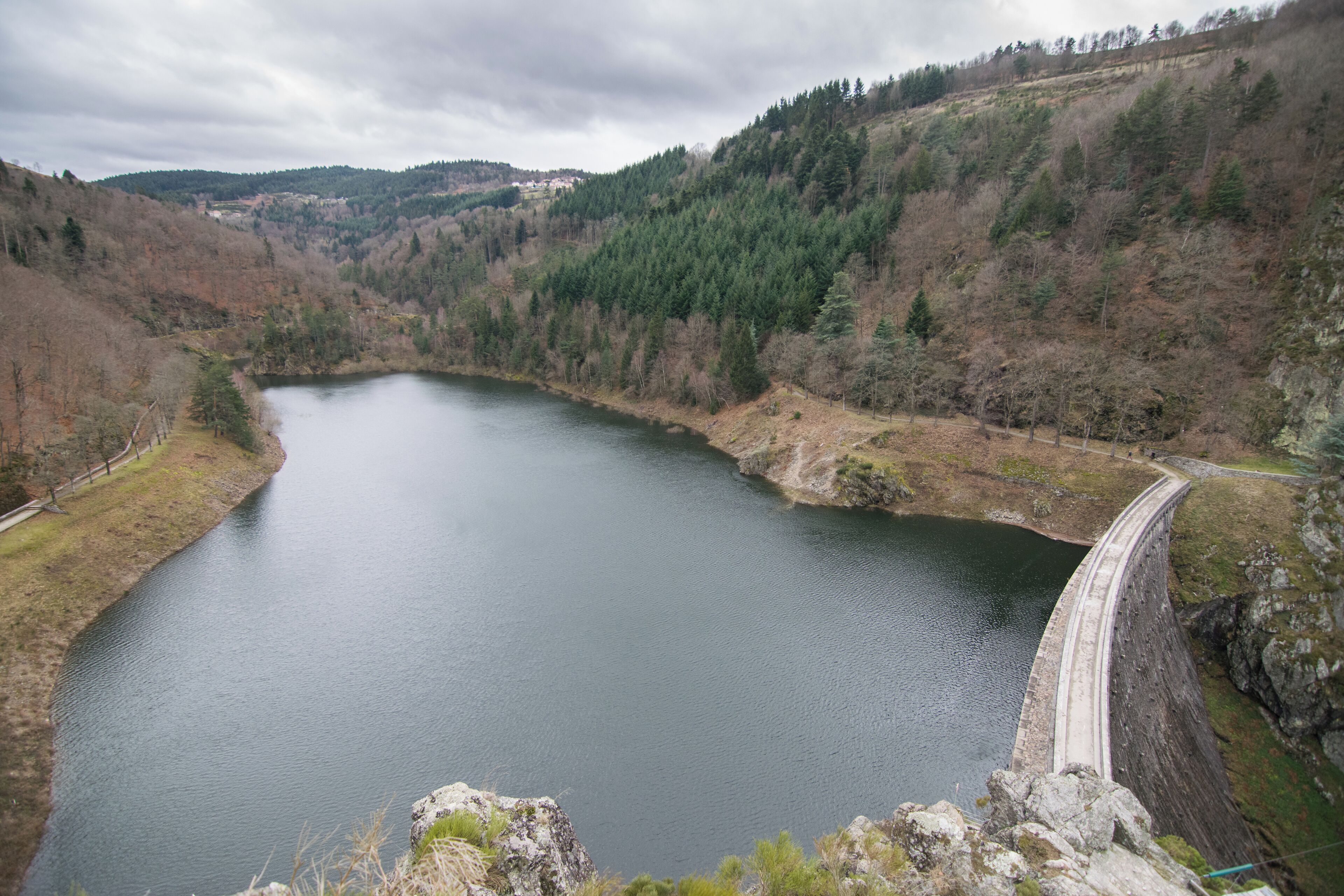 A large reservoir with a dam, surrounded by forested hills and a cloudy sky.