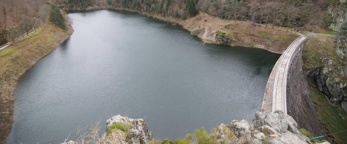 A large reservoir with a dam, surrounded by forested hills and a cloudy sky.