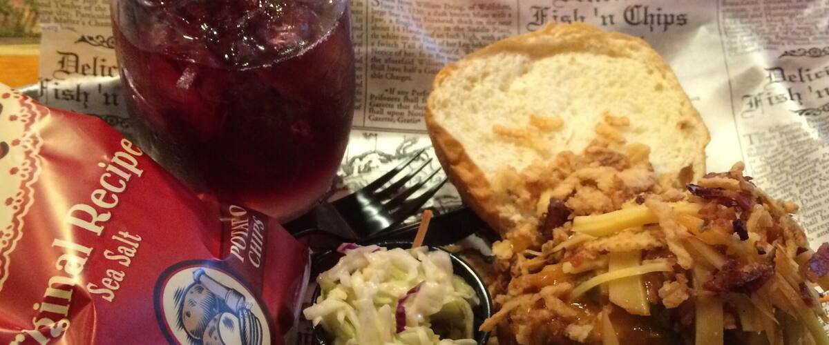 A meal with a sandwich, coleslaw, chips, and a drink on a newspaper.