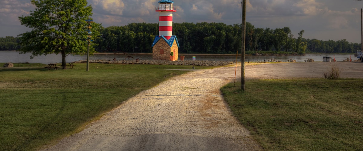 A gravel path leading to a red and white striped lighthouse with a blue roof, surrounded by trees and a body of water.