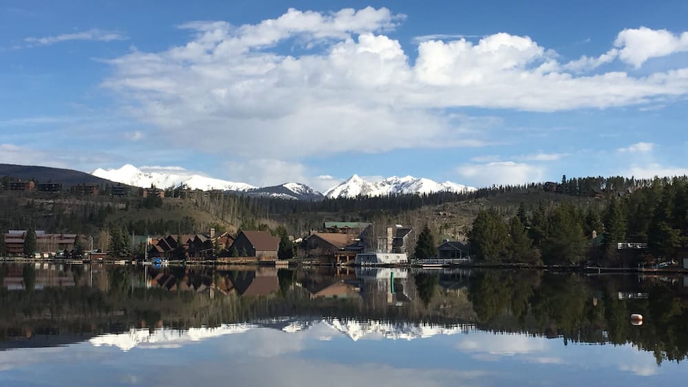 Ein ruhiger See spiegelt Gebäude und schneebedeckte Berge unter einem teils bewölkten Himmel wider.