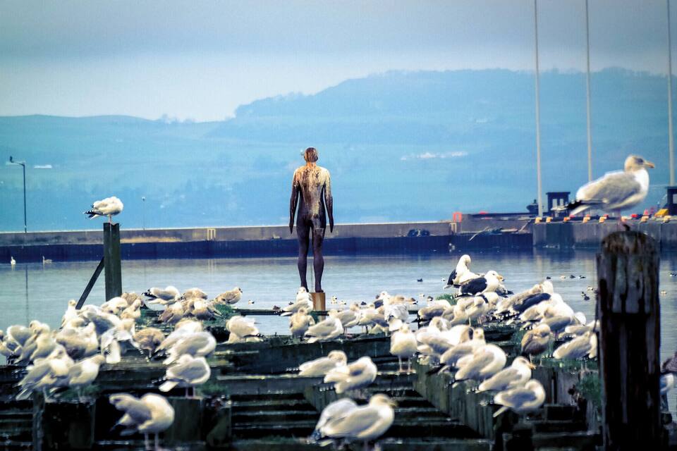 Une statue représentant une figure humaine, placée sur une jetée et entourée de mouettes.