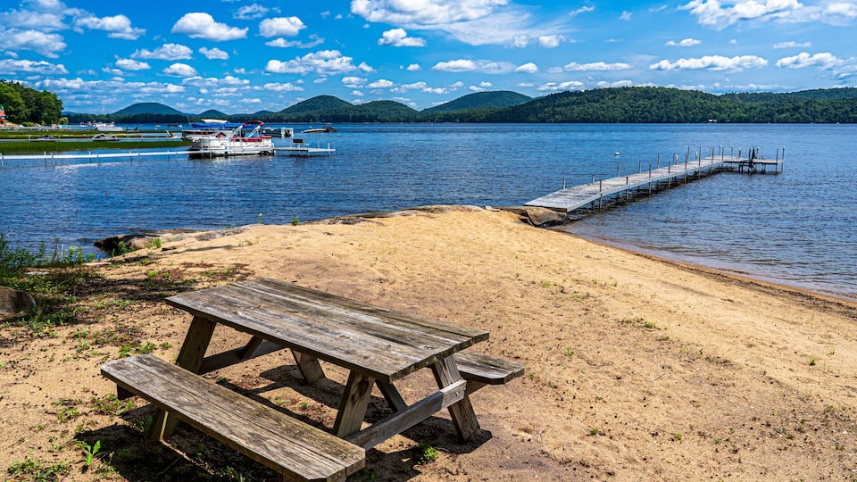 A sandy beach with a wooden picnic table and a dock extending into a lake with mountains in the background.