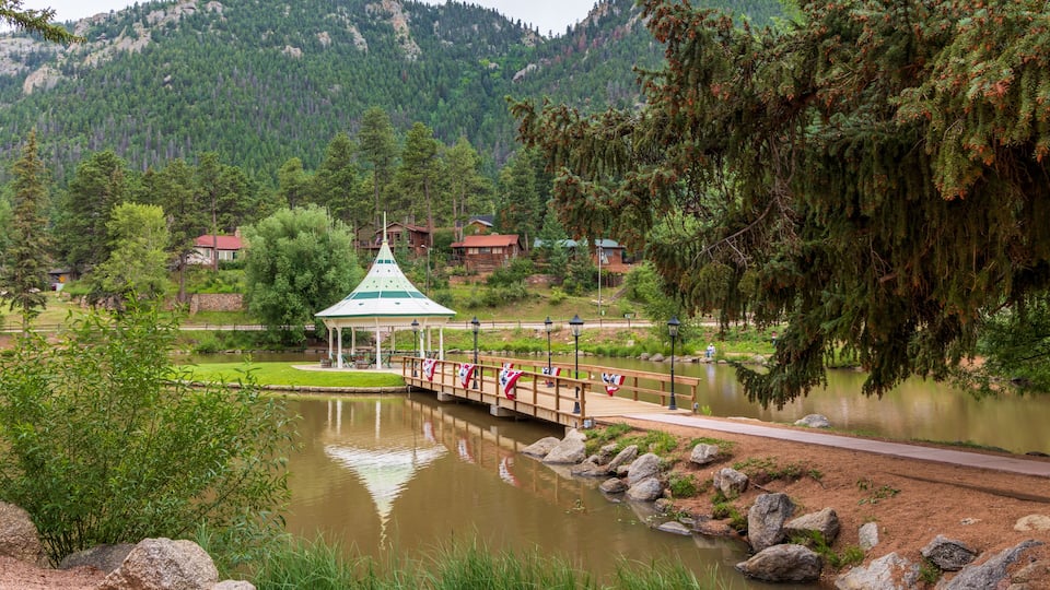 A tranquil lake with a wooden bridge, a gazebo, and houses in a mountainous backdrop.