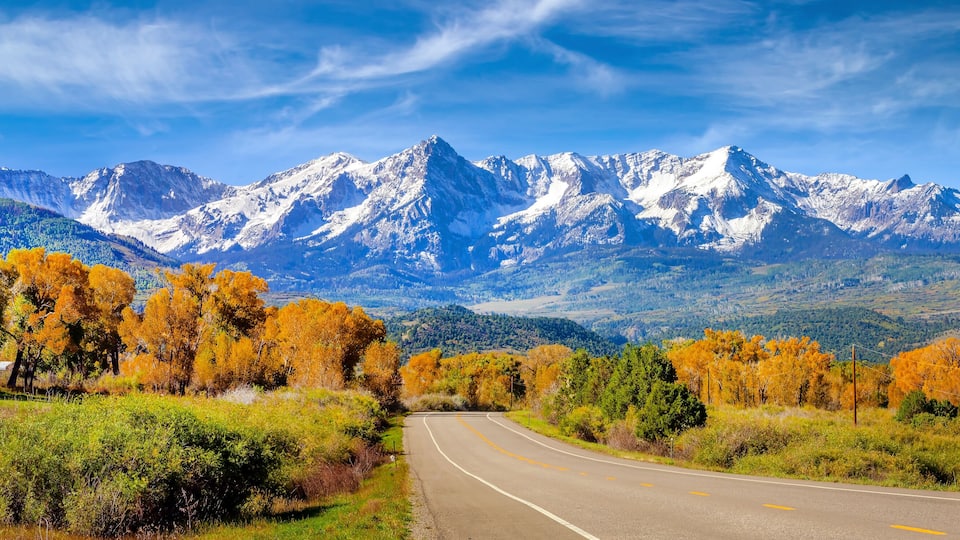 A winding road with autumn foliage, a mountain range with snow-capped peaks, and a clear blue sky.