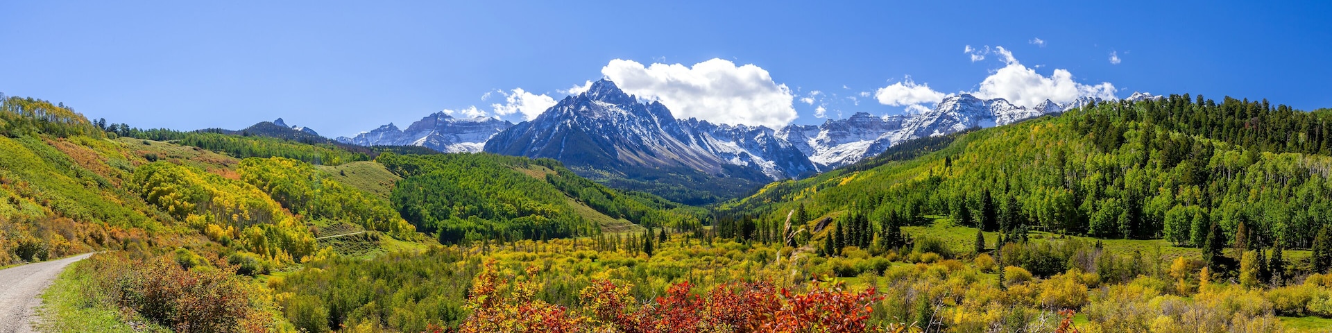 A mountain range with a clear sky, a dirt road, and autumn-colored foliage.