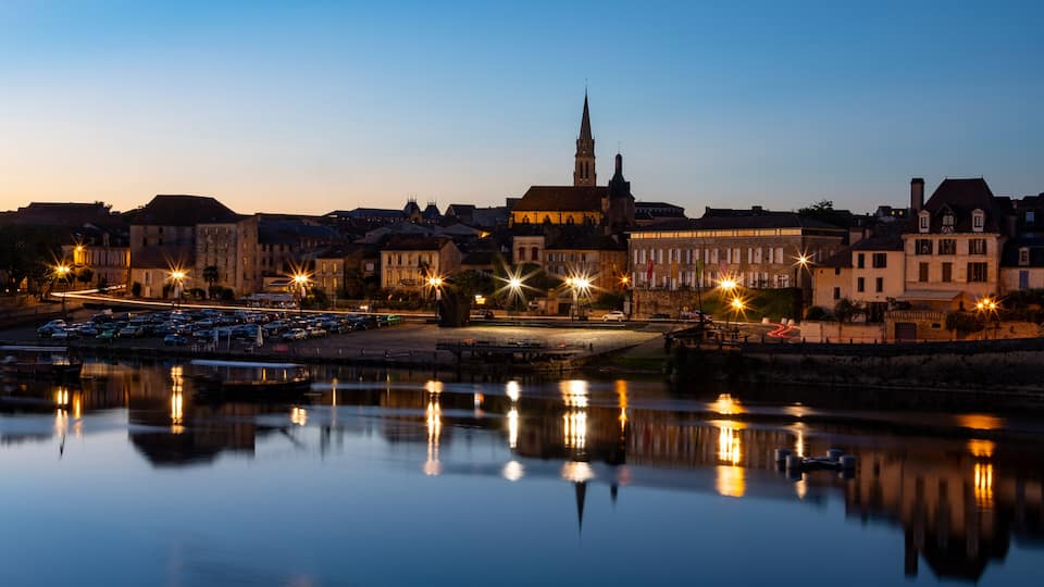 A cityscape at dusk with a prominent church spire, a calm river reflecting lights, and a parking lot with cars.