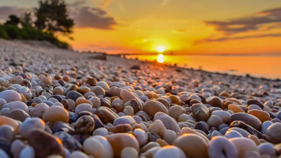 A pebbled beach at sunset with the sun setting over the horizon.