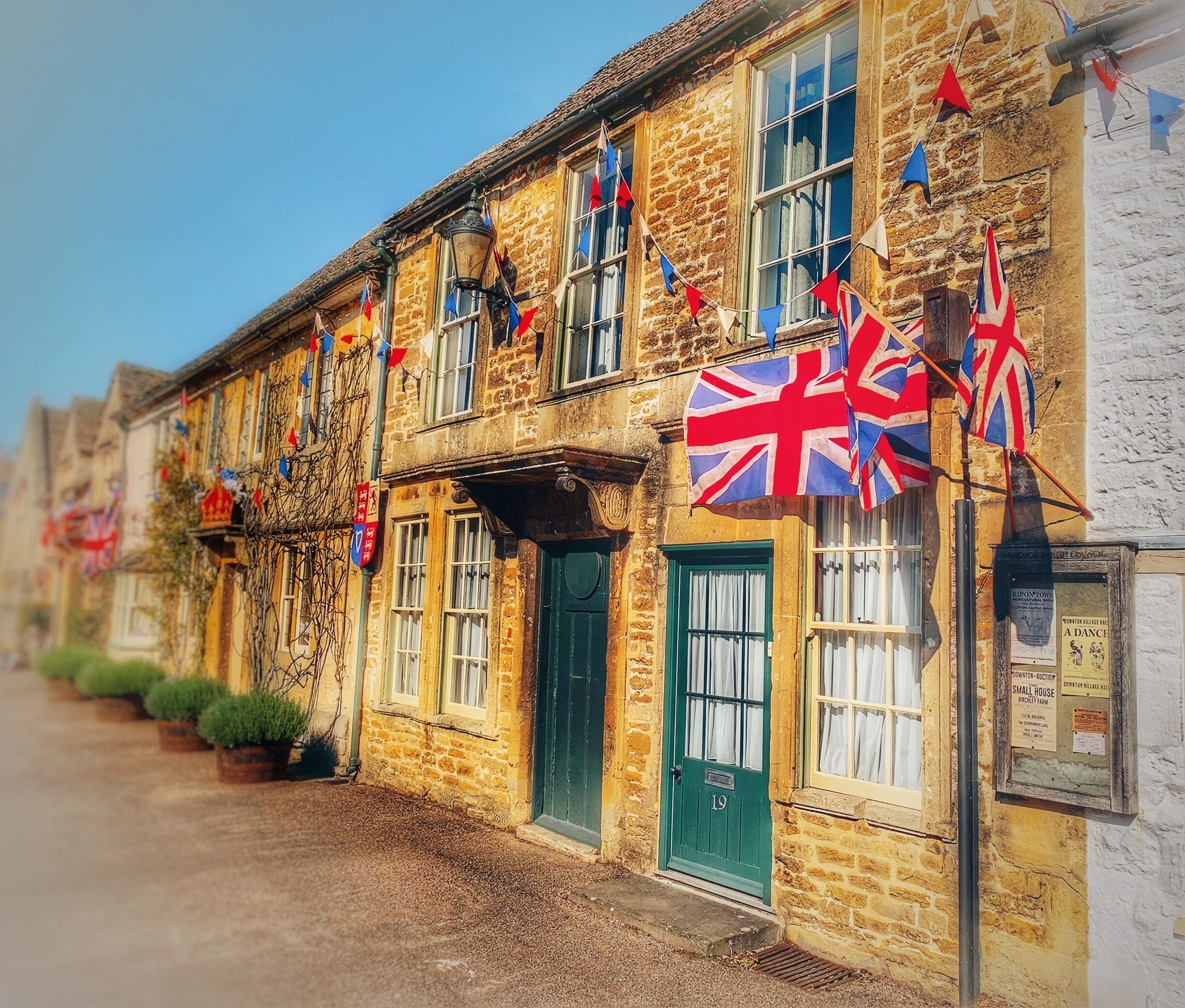A row of stone buildings with British flags and a street lined with potted plants.