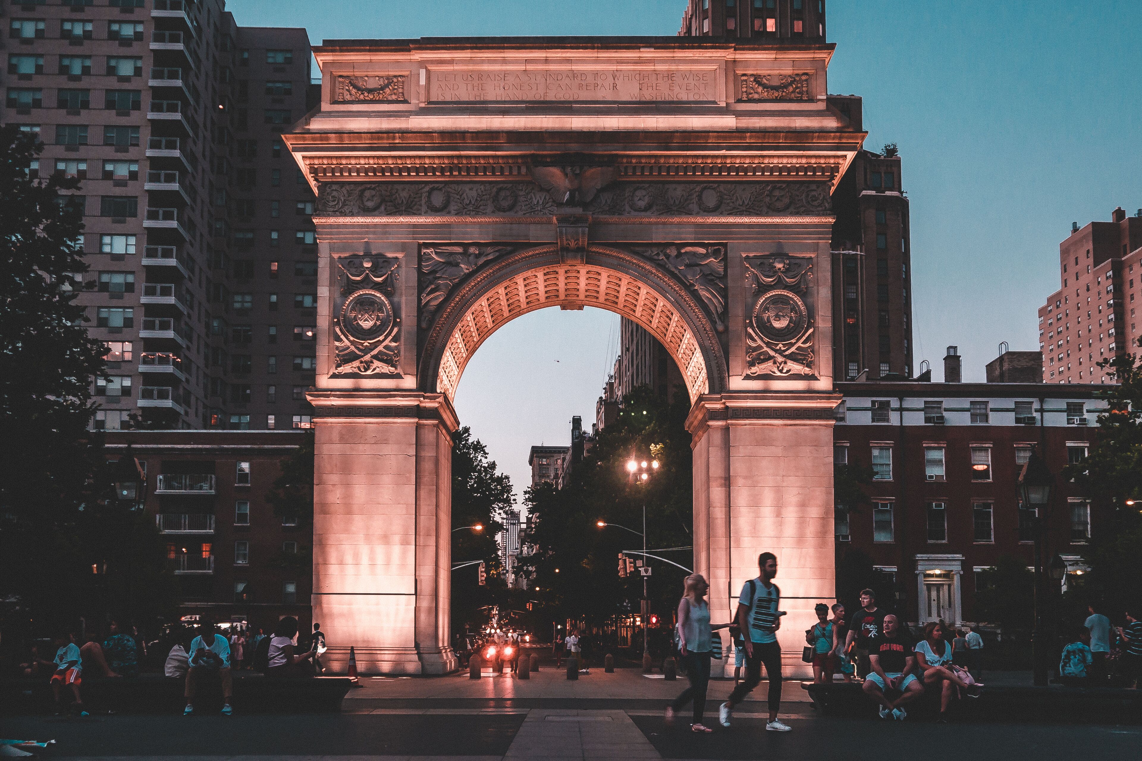 An illuminated archway with intricate carvings, surrounded by urban buildings and people.