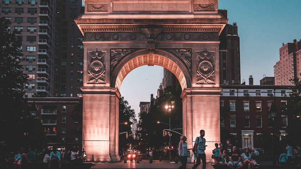 An illuminated archway with intricate carvings, surrounded by urban buildings and people.