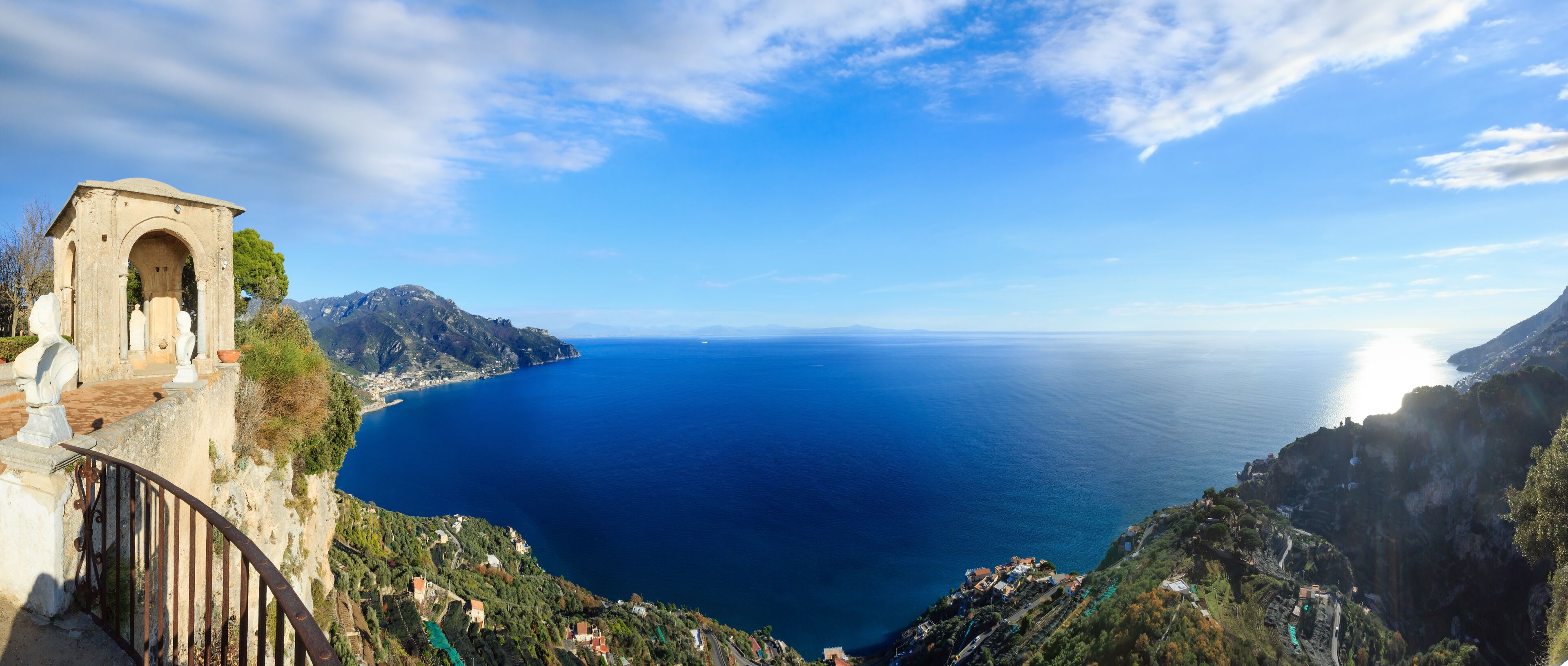 A coastal landscape with a stone structure, a clear blue sea, and mountainous terrain.