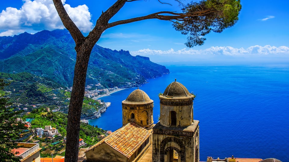 A coastal town with two prominent church towers, lush green hills, and a clear blue sea.