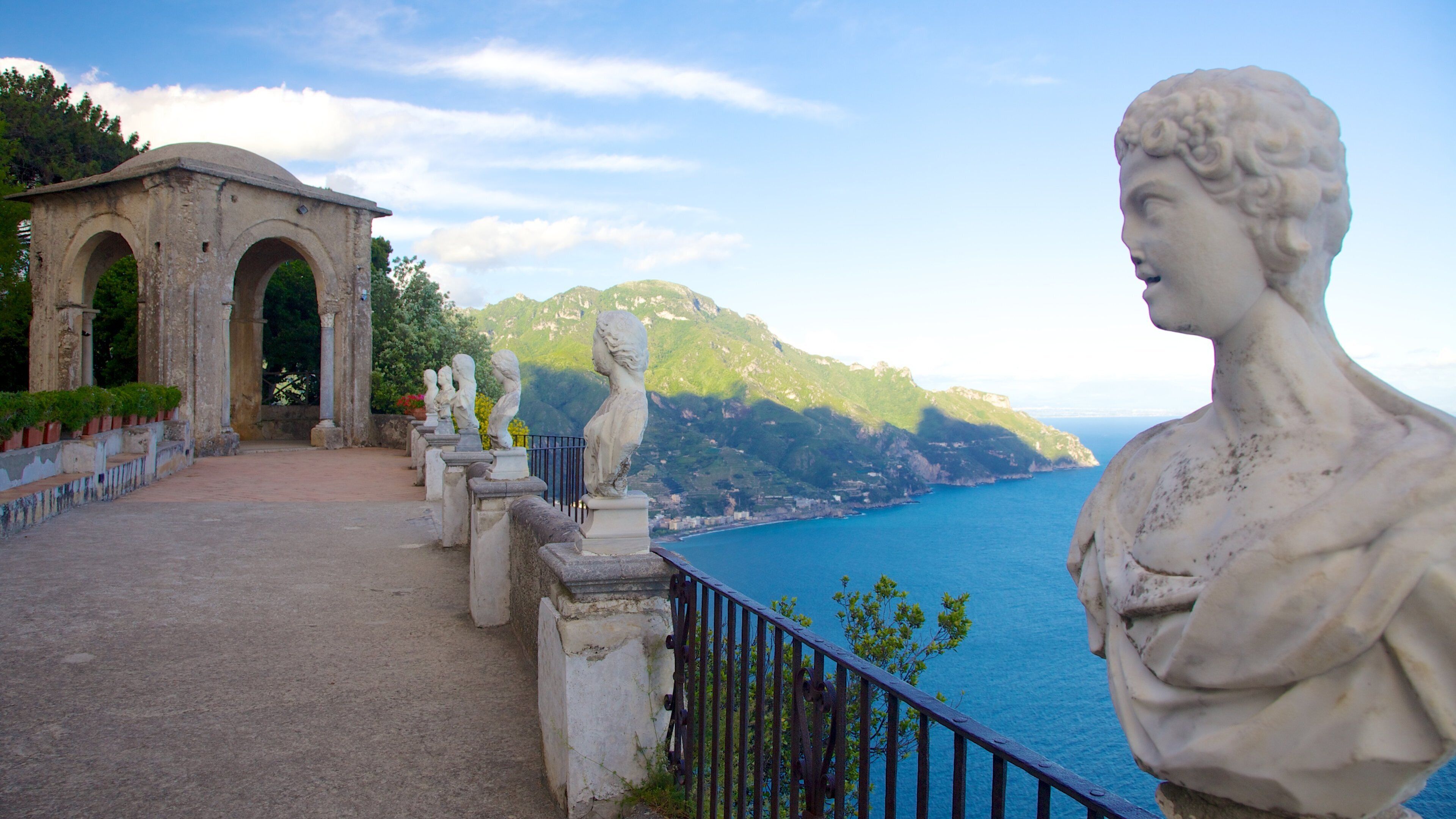 A coastal landscape with a statue, a stone archway, and a view of the sea and mountains.