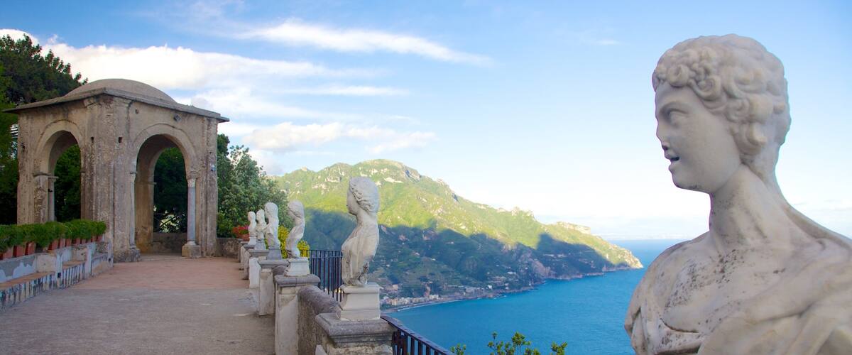 A coastal landscape with a statue, a stone archway, and a view of the sea and mountains.