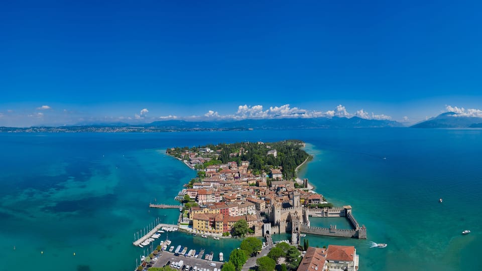 An aerial view of a coastal town with a historic castle, clear blue waters, and distant mountains.
