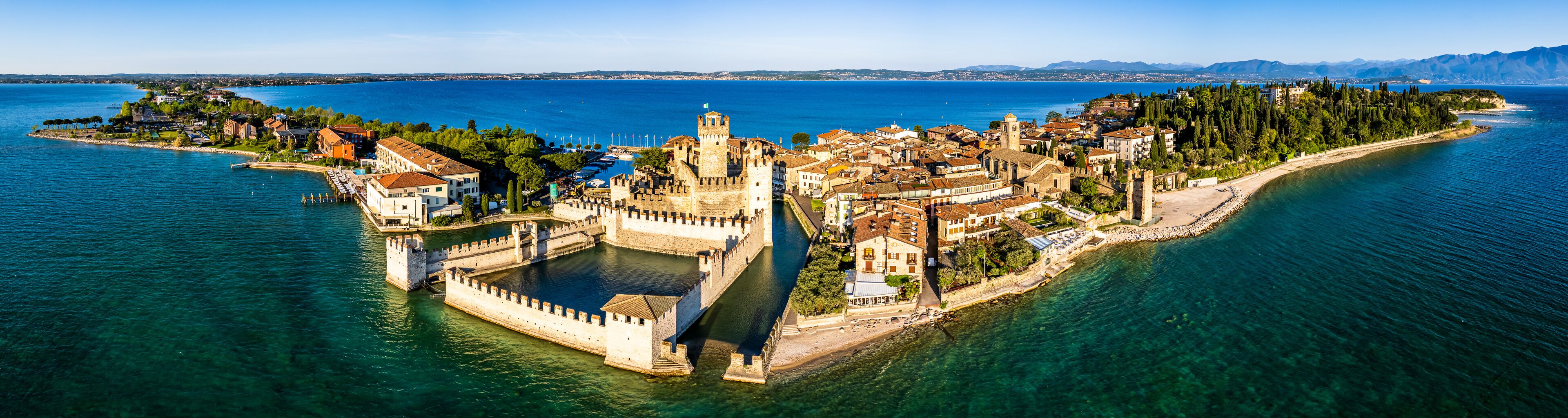 An aerial view of a coastal town with a prominent castle, clear blue sea, and lush greenery.