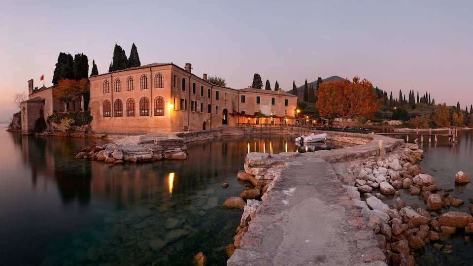 A historic building with a stone pathway leading to a calm lake, surrounded by trees and a distant mountain.