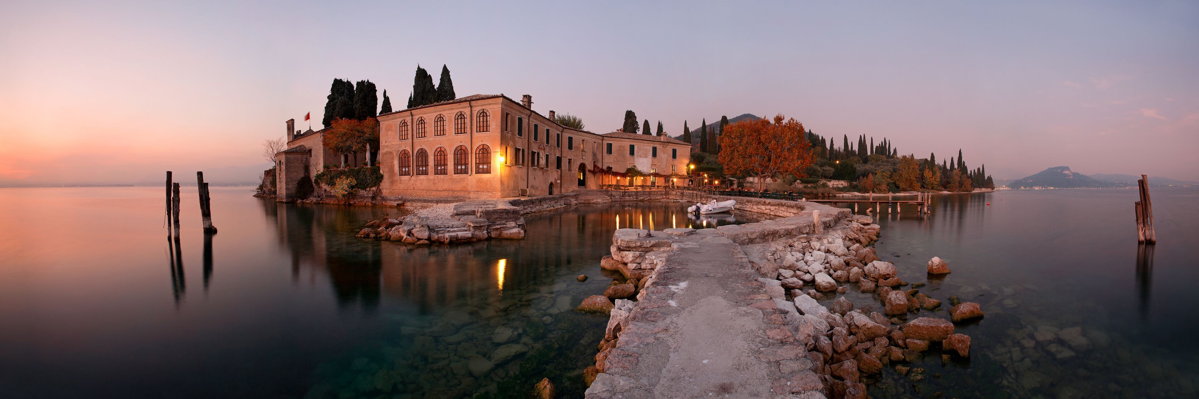 A historic building with a stone pathway leading to a calm lake, surrounded by trees and a distant mountain.