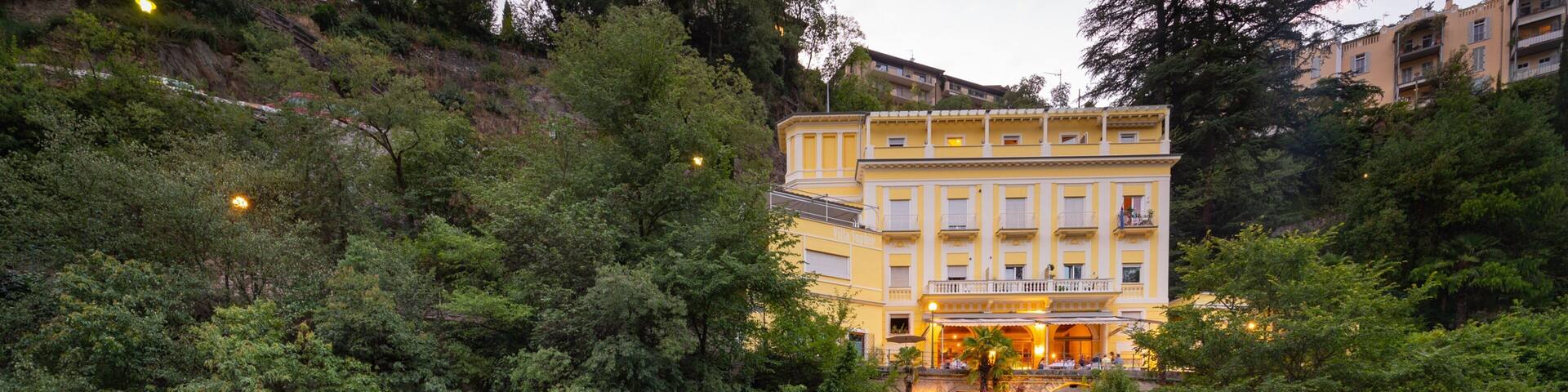 A river with rocks, surrounded by trees and a yellow building with balconies.