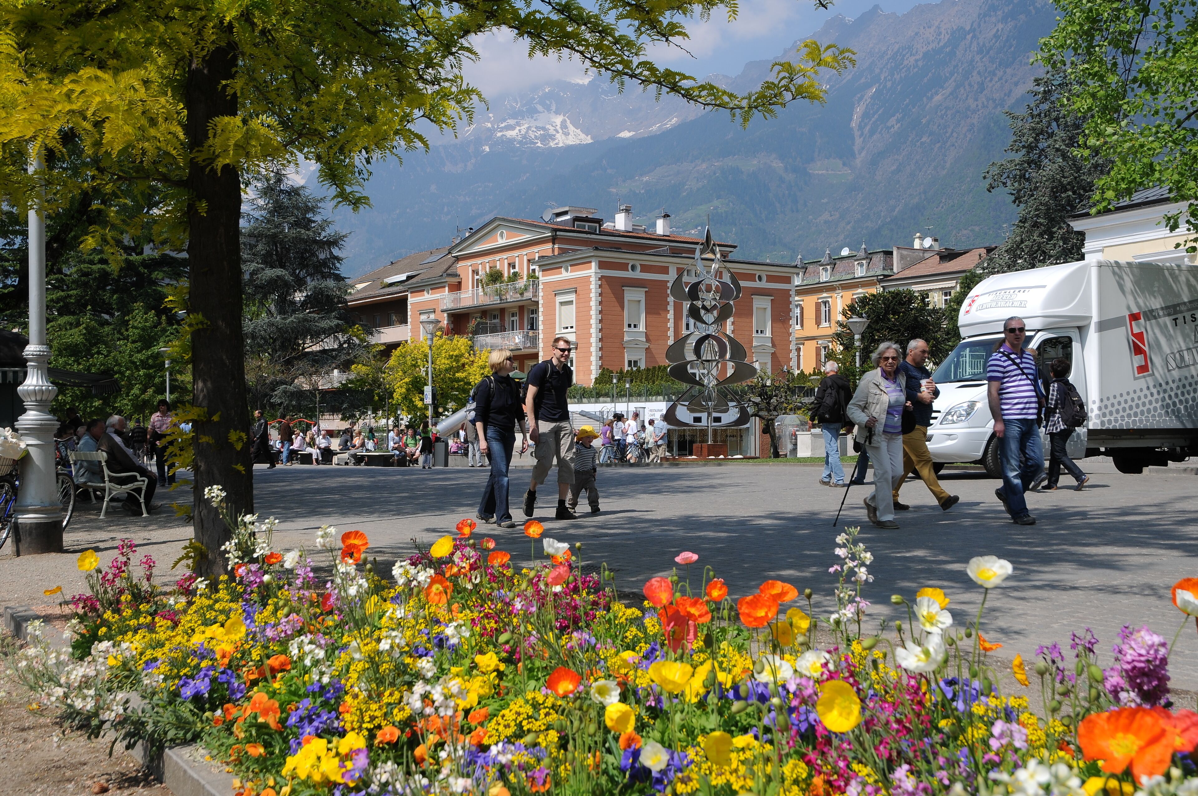 Uma praça na cidade com flores coloridas, um edifício grande com um relógio e montanhas ao fundo.