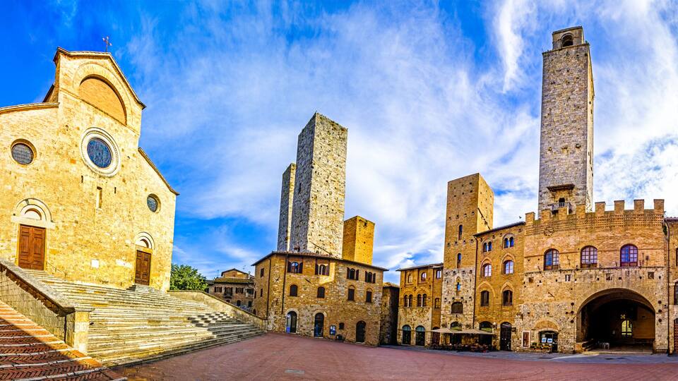 A historic town square with medieval architecture, including a church with a bell tower and a tall, narrow tower.