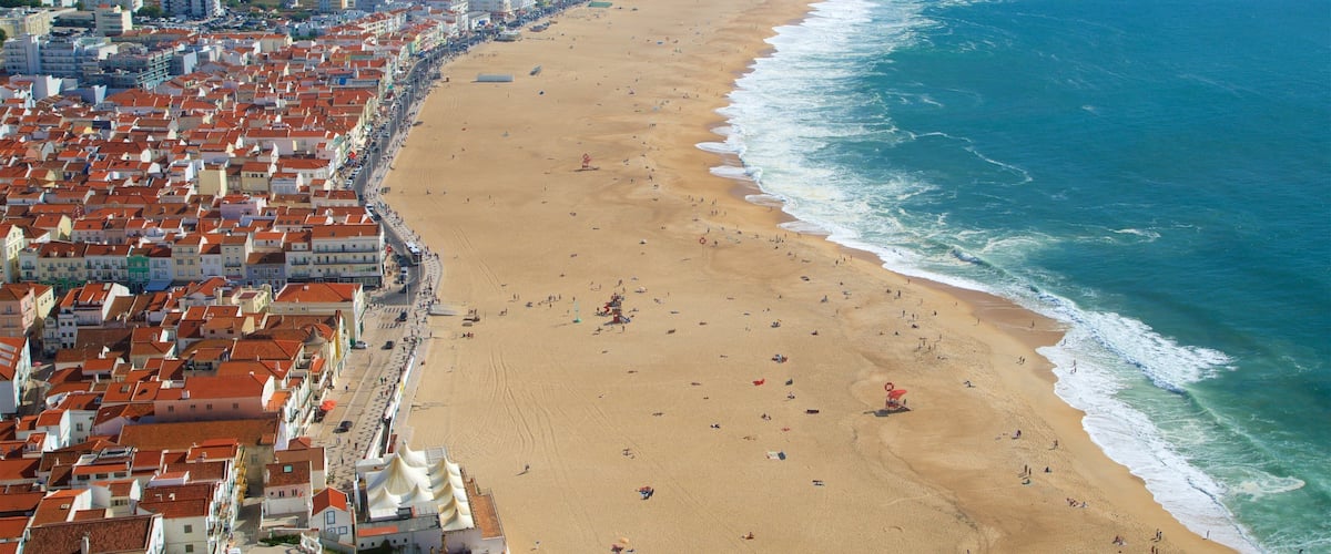 A coastal city with a wide sandy beach, a dense cluster of red-roofed buildings, and a clear view of the ocean.