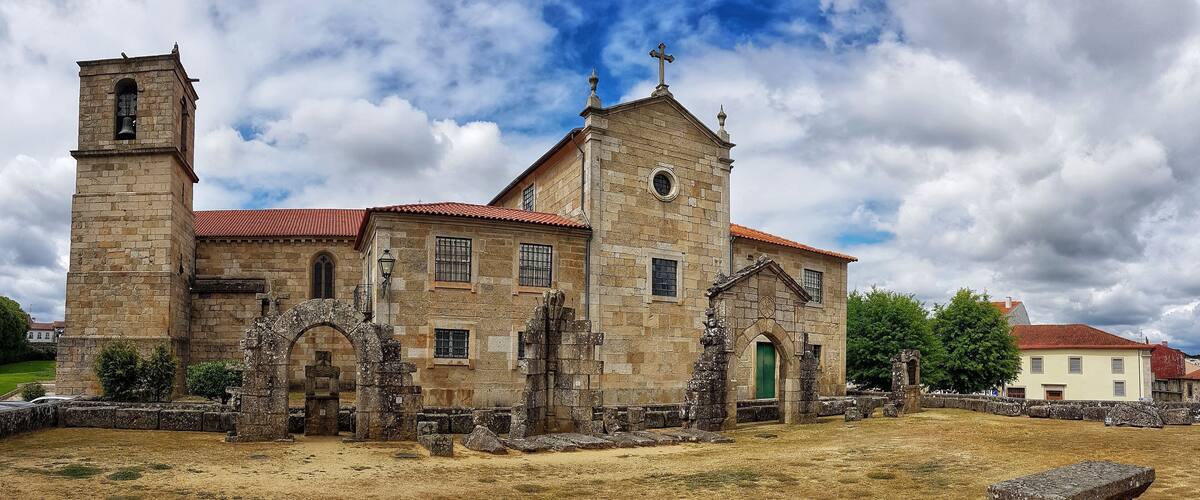 A historic stone church with a bell tower and a cross on top, surrounded by ruins and a dry field.