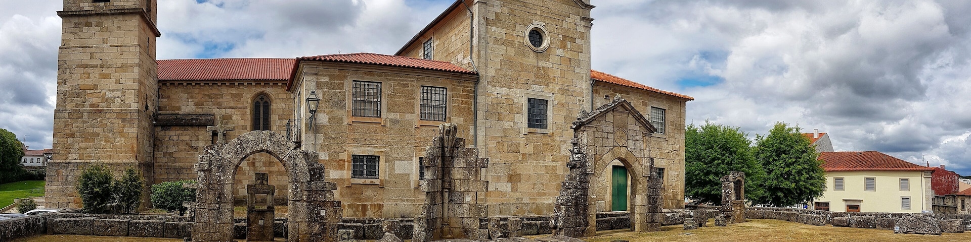 A historic stone church with a bell tower and a cross on top, surrounded by ruins and a dry field.