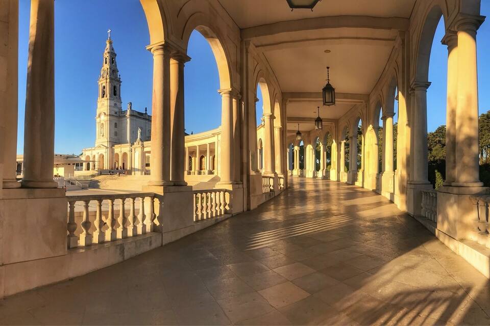 A covered walkway with columns and arches, leading to a church with a tall bell tower.
