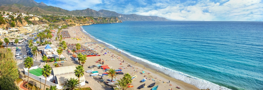 A coastal town with a beach, parked cars, and palm trees.
