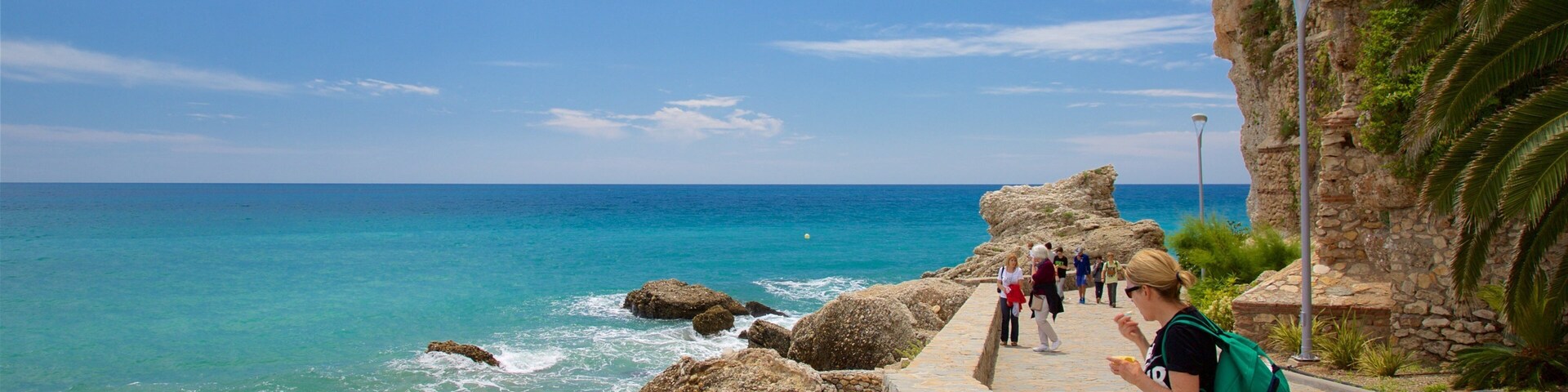 Un paseo marítimo con rocas grandes, un mar azul cristalino y un edificio con balcón con vistas al océano.