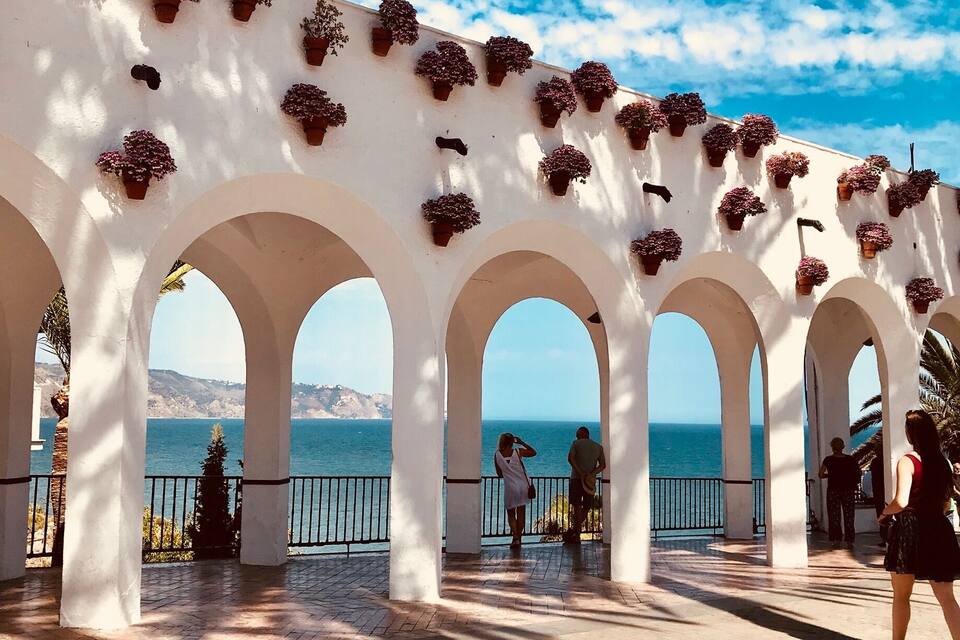 A white building with arches and potted plants, overlooking a body of water.