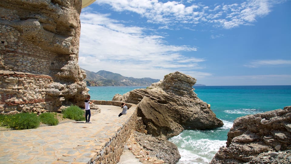 A coastal stone pathway leading to a rocky outcrop by the sea, with a clear sky and distant mountains.