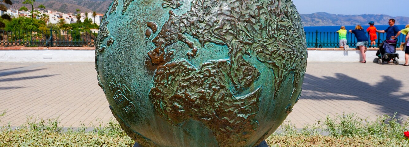 Una escultura de bronce de un globo terráqueo con inscripciones, sobre un pedestal, con un cielo azul claro y montañas al fondo.