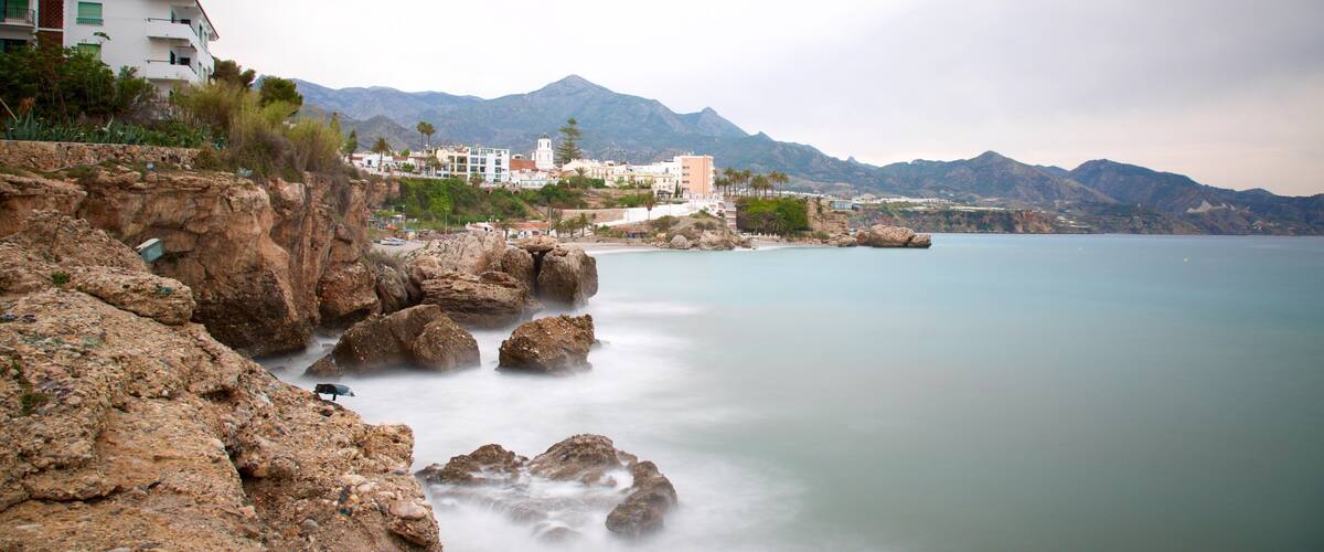 A coastal landscape with rocky cliffs, calm sea, and buildings in the background.
