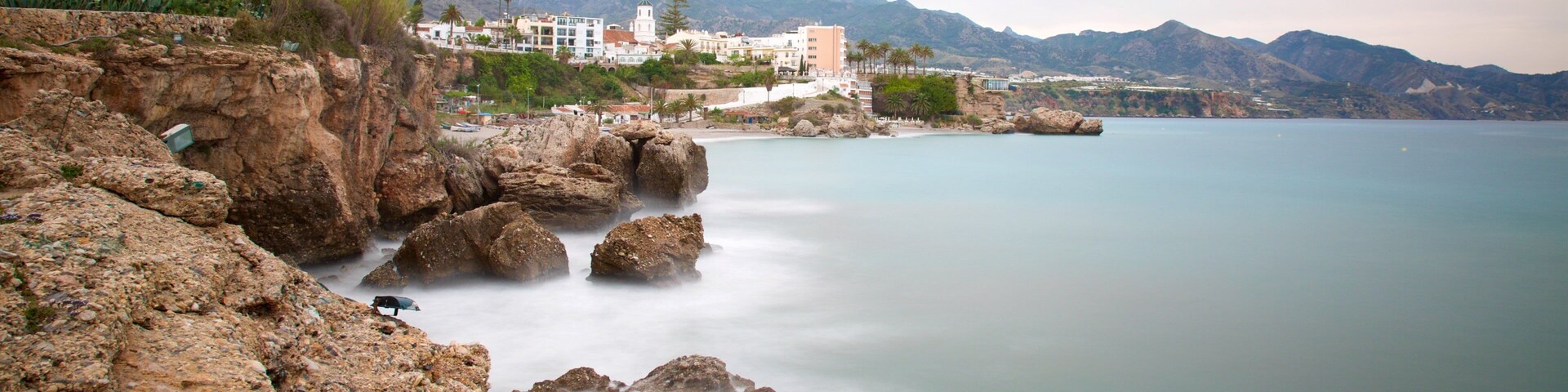 A coastal landscape with rocky cliffs, calm sea, and buildings in the background.