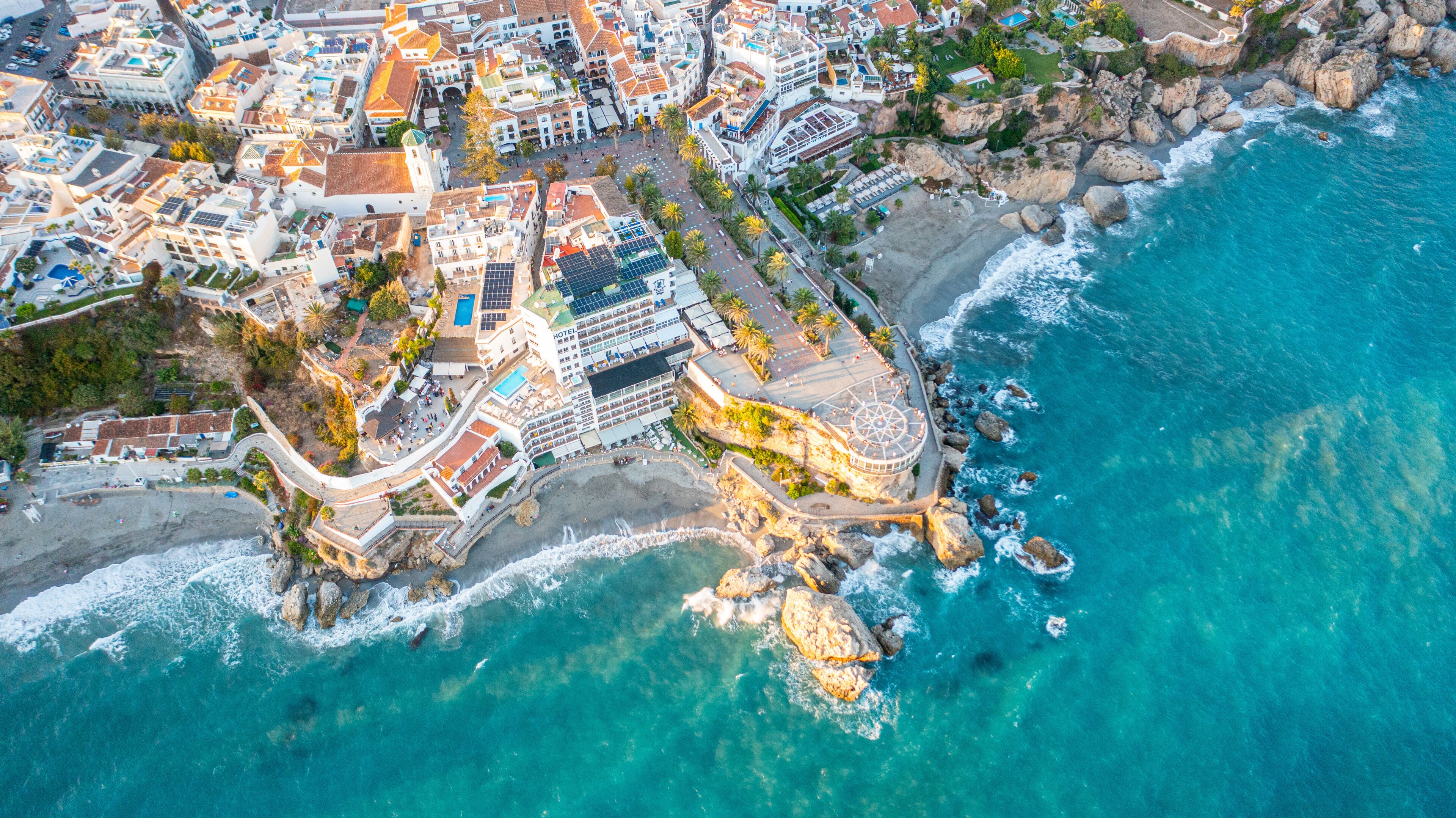 An aerial view of a coastal town with a prominent hotel, rocky shoreline, and clear blue waters.