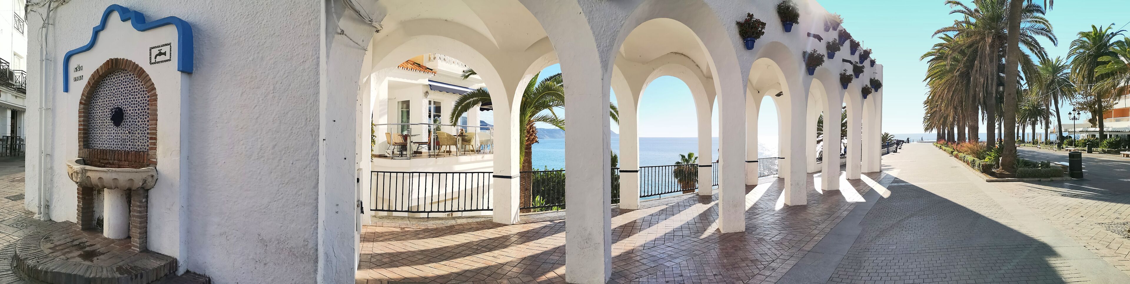 A coastal promenade with white arches, a view of the sea, and palm trees.